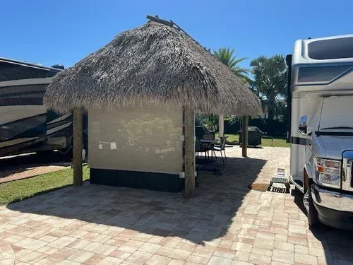Tiki hut with tan woven roof and shade screens next to a parked RV on a brick patio.