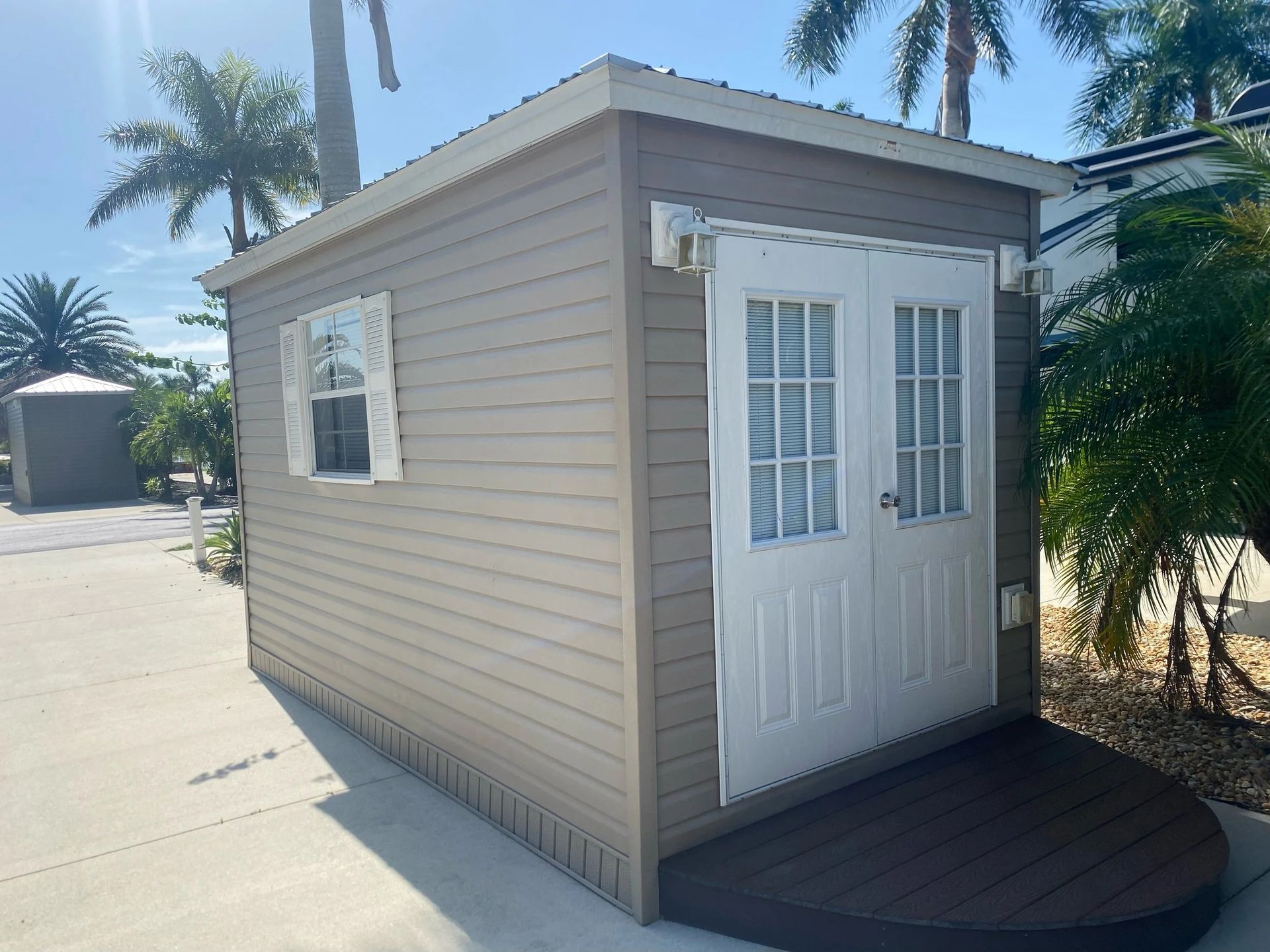 Small tan shed with white double doors, small window, and a dark brown deck; palm trees in background.