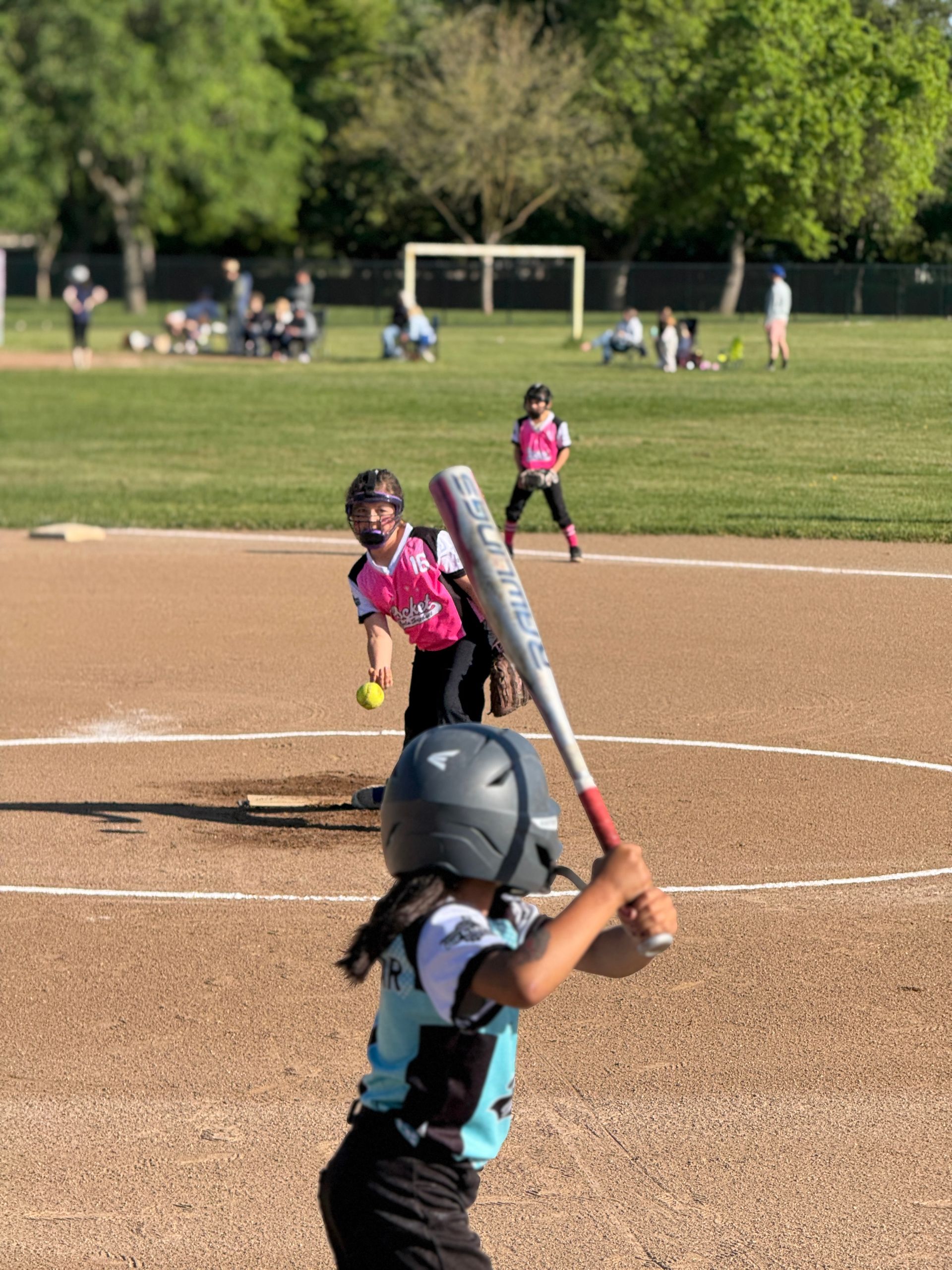 A person wearing a hat and orange shirt, holding a baseball bat on their shoulder on a baseball field.