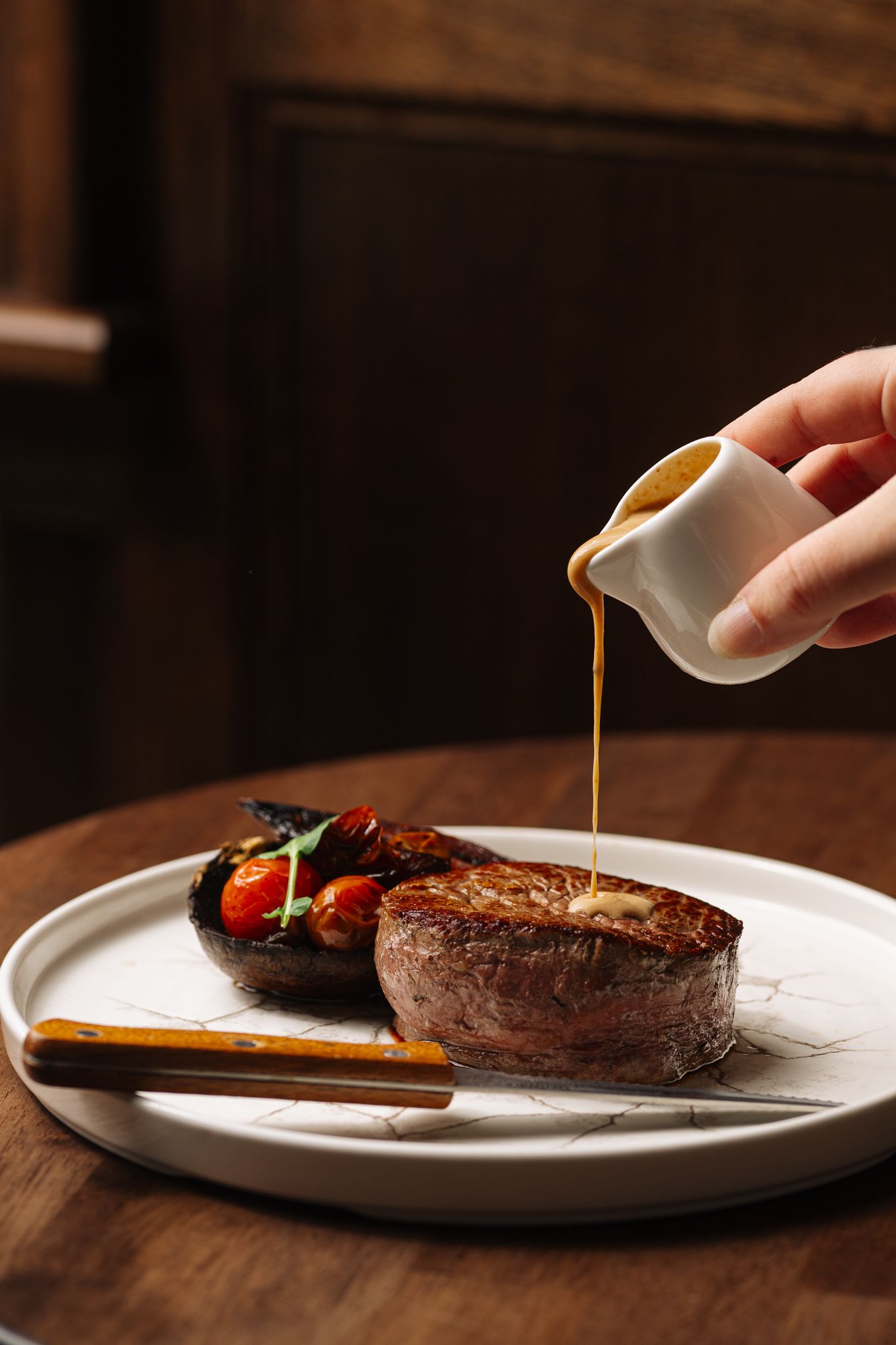 A person is pouring sauce on a steak on a plate.