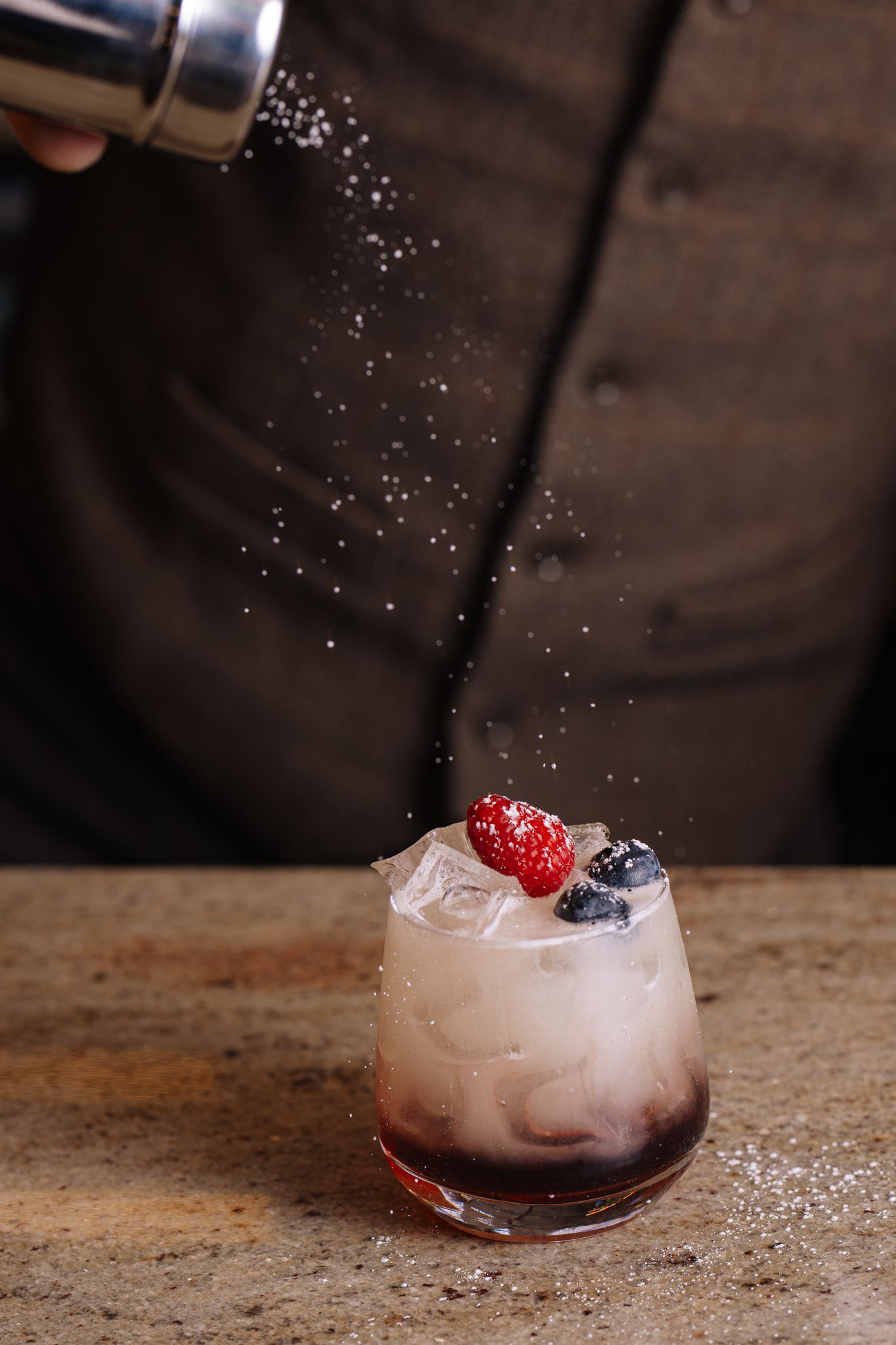 A person is pouring sugar into a drink with berries on top.
