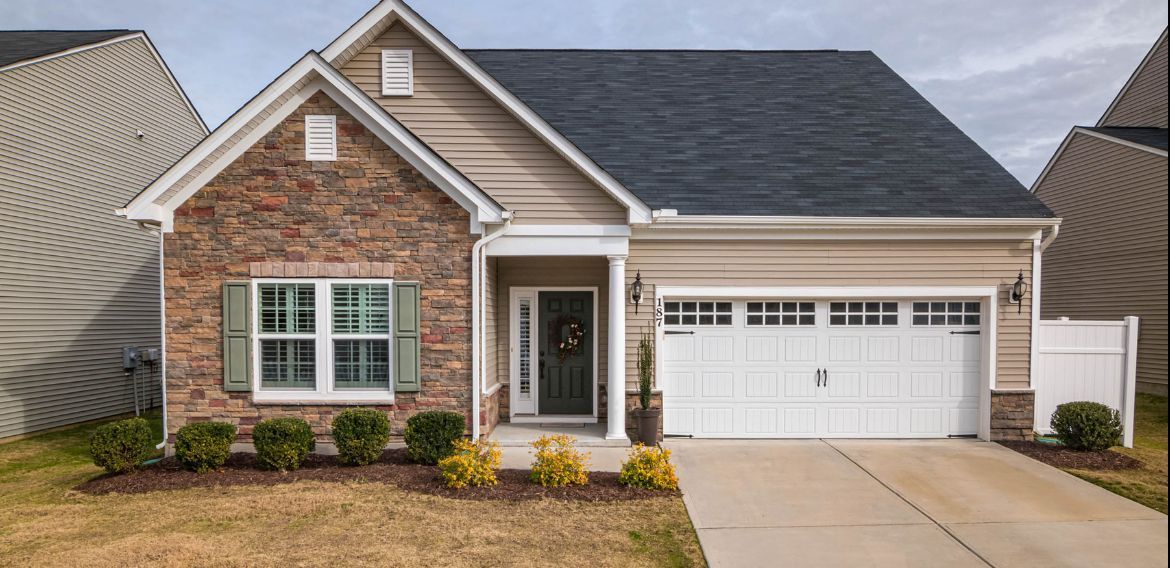 Tan and stone fronted house with a black door, white garage door, and green bushes.