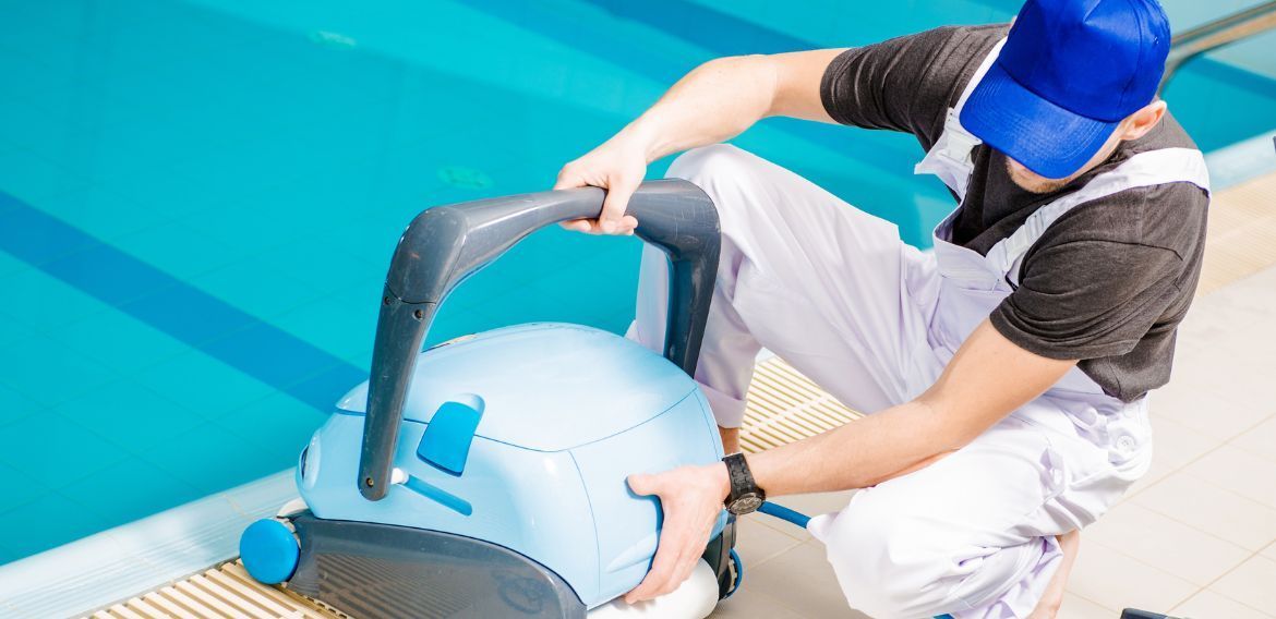 Person in blue cap and white overalls tending to a robotic pool cleaner at the edge of a pool.