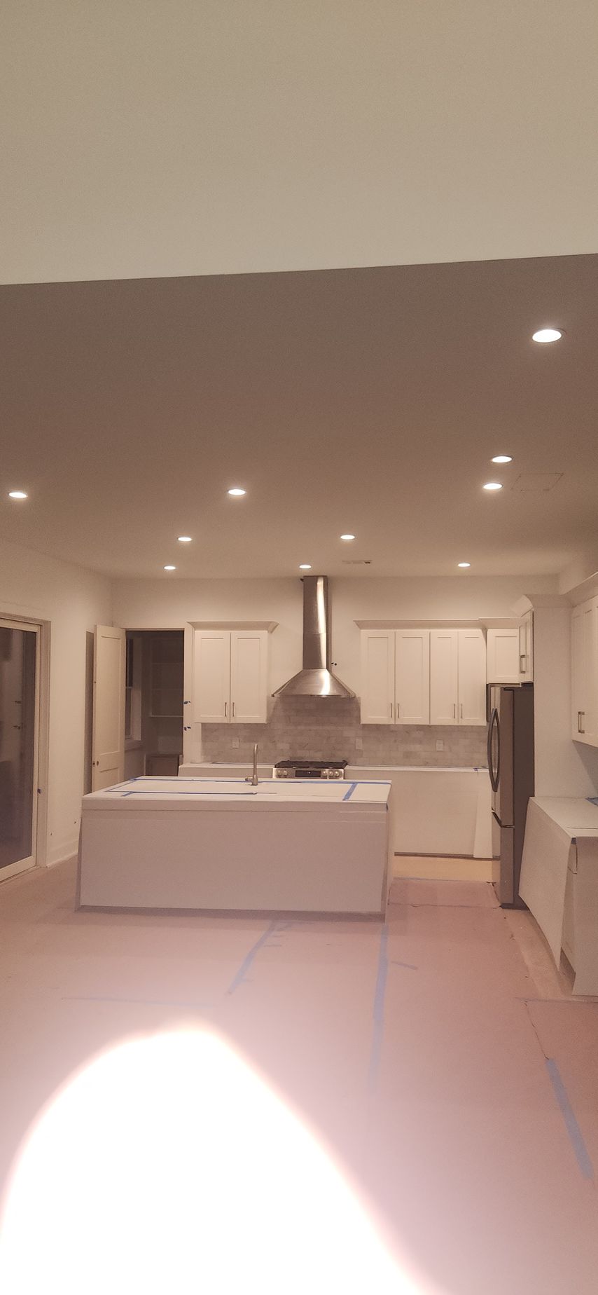 A modern, white kitchen with a stainless steel range hood, island, and recessed lighting. Pink floor covering.