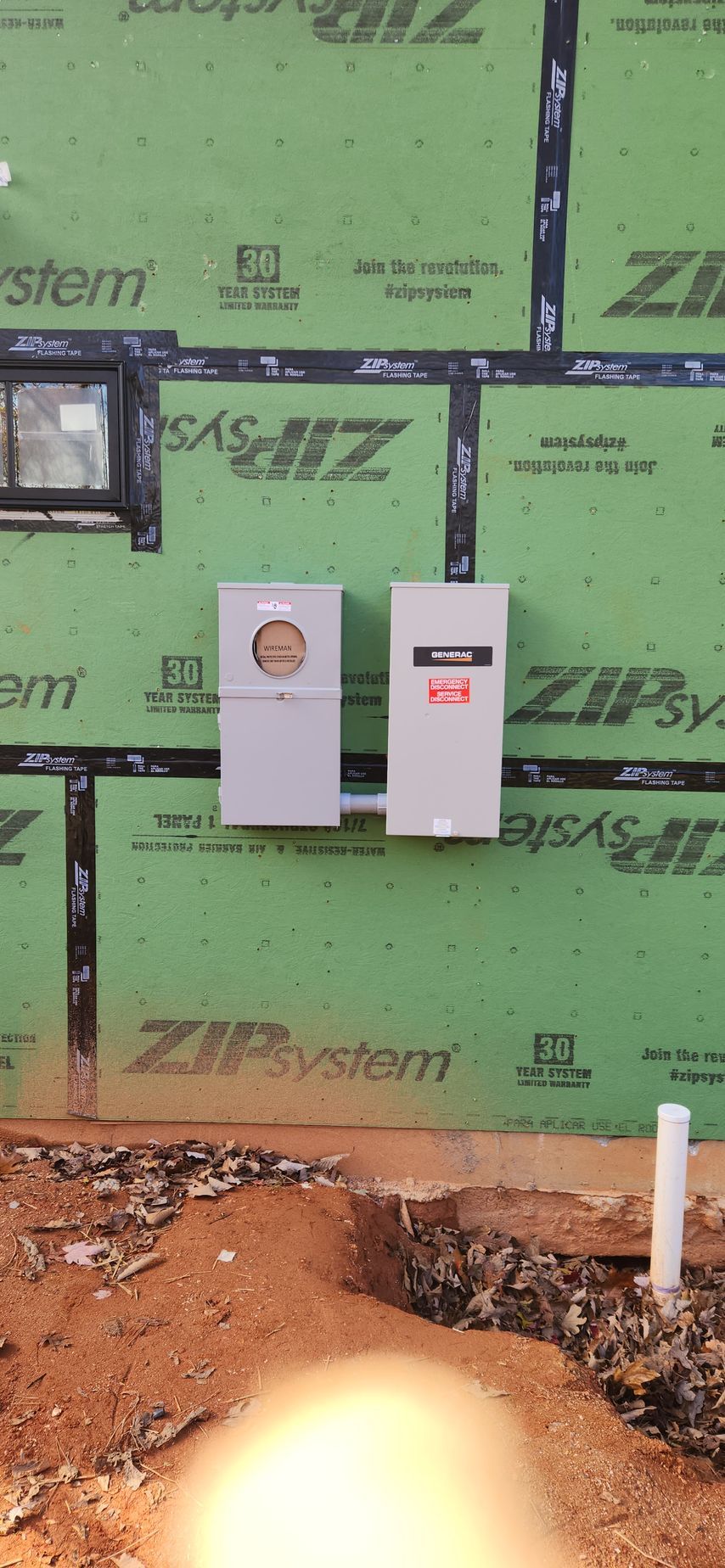 Electrical meter and panel on a green Zip System wall. Construction site with exposed dirt and a white pipe.