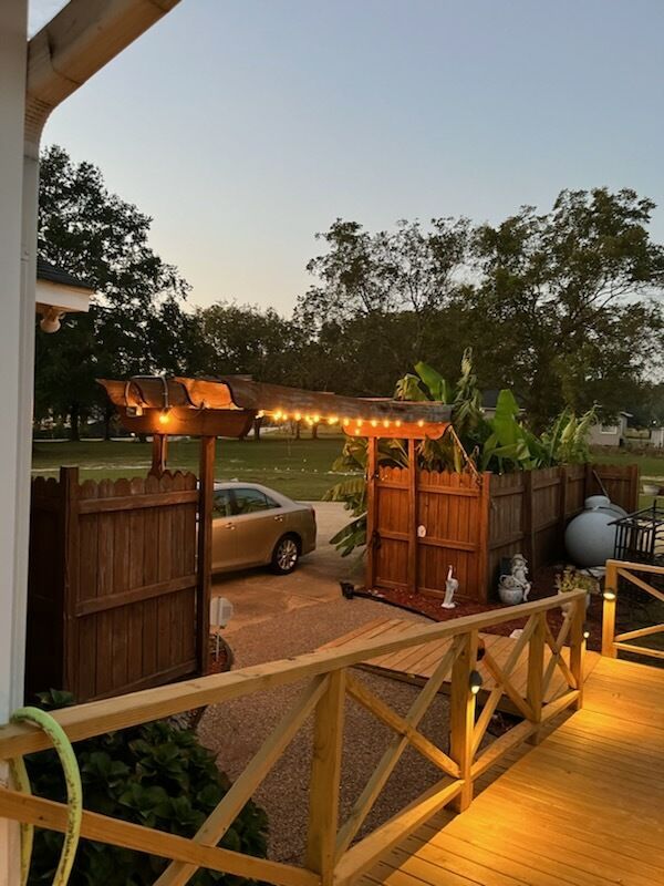 Wooden deck with railing leading to a courtyard with a pergola and string lights. A car is parked nearby.