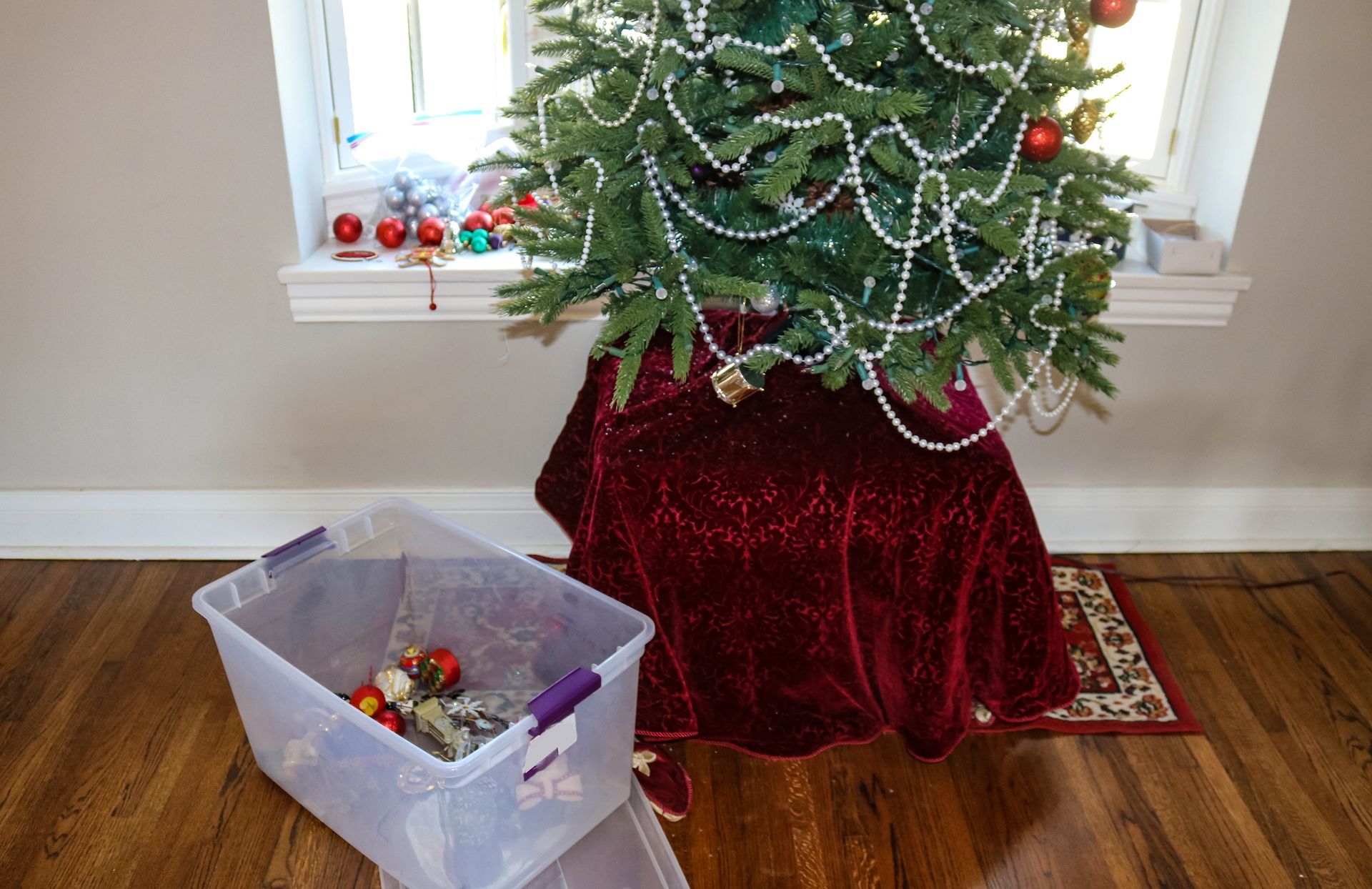 Christmas tree skirted in red velvet, partially decorated, with a storage bin of ornaments nearby.