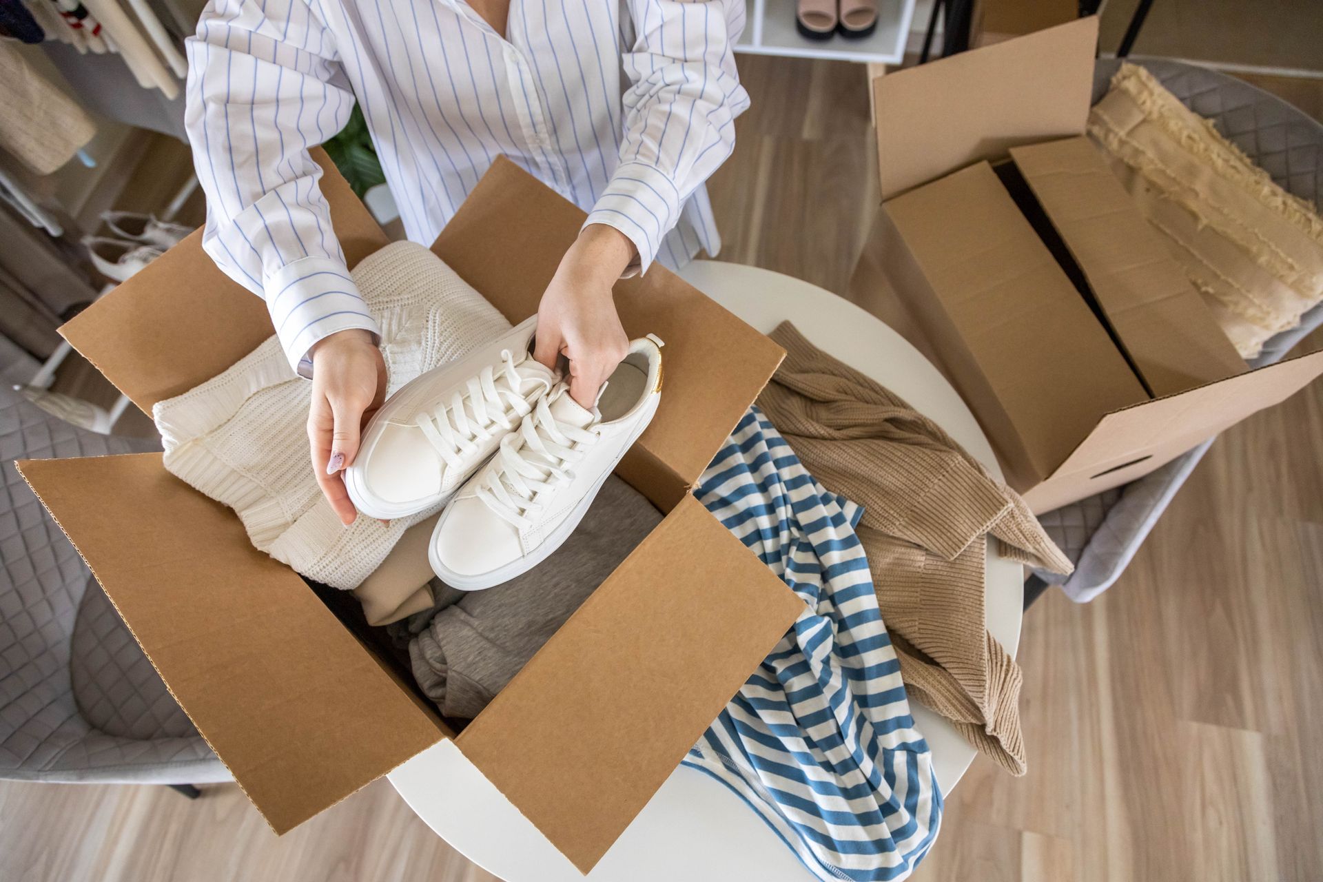 Woman packing a cardboard box with clothes and shoes on a white table.
