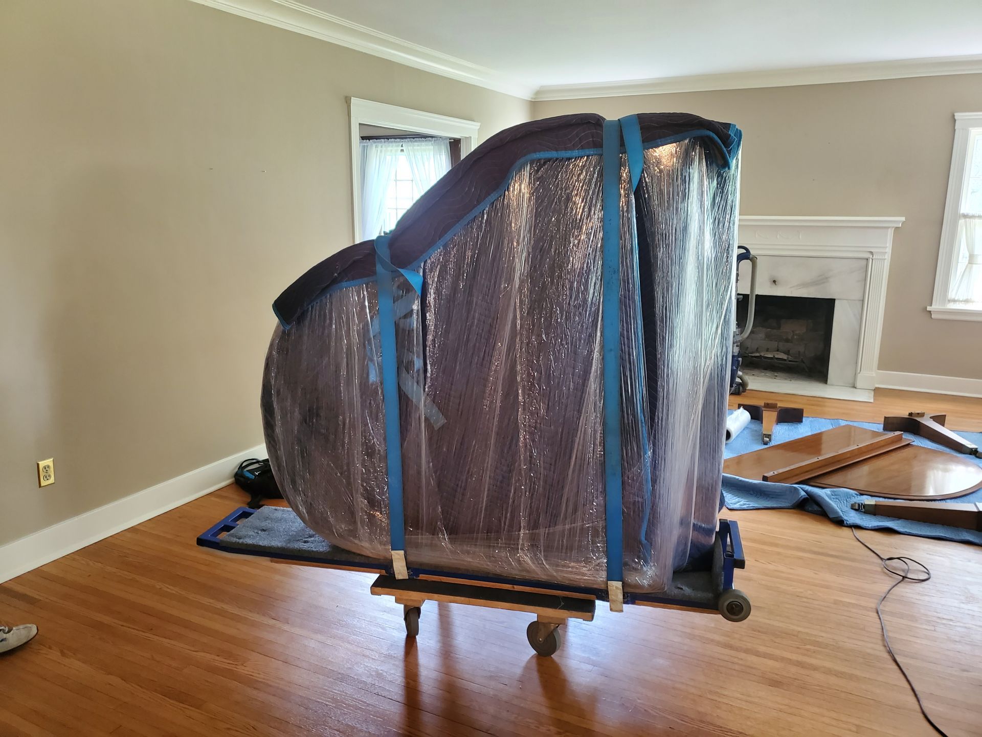 A piano is wrapped in plastic and sitting on a cart in a living room.