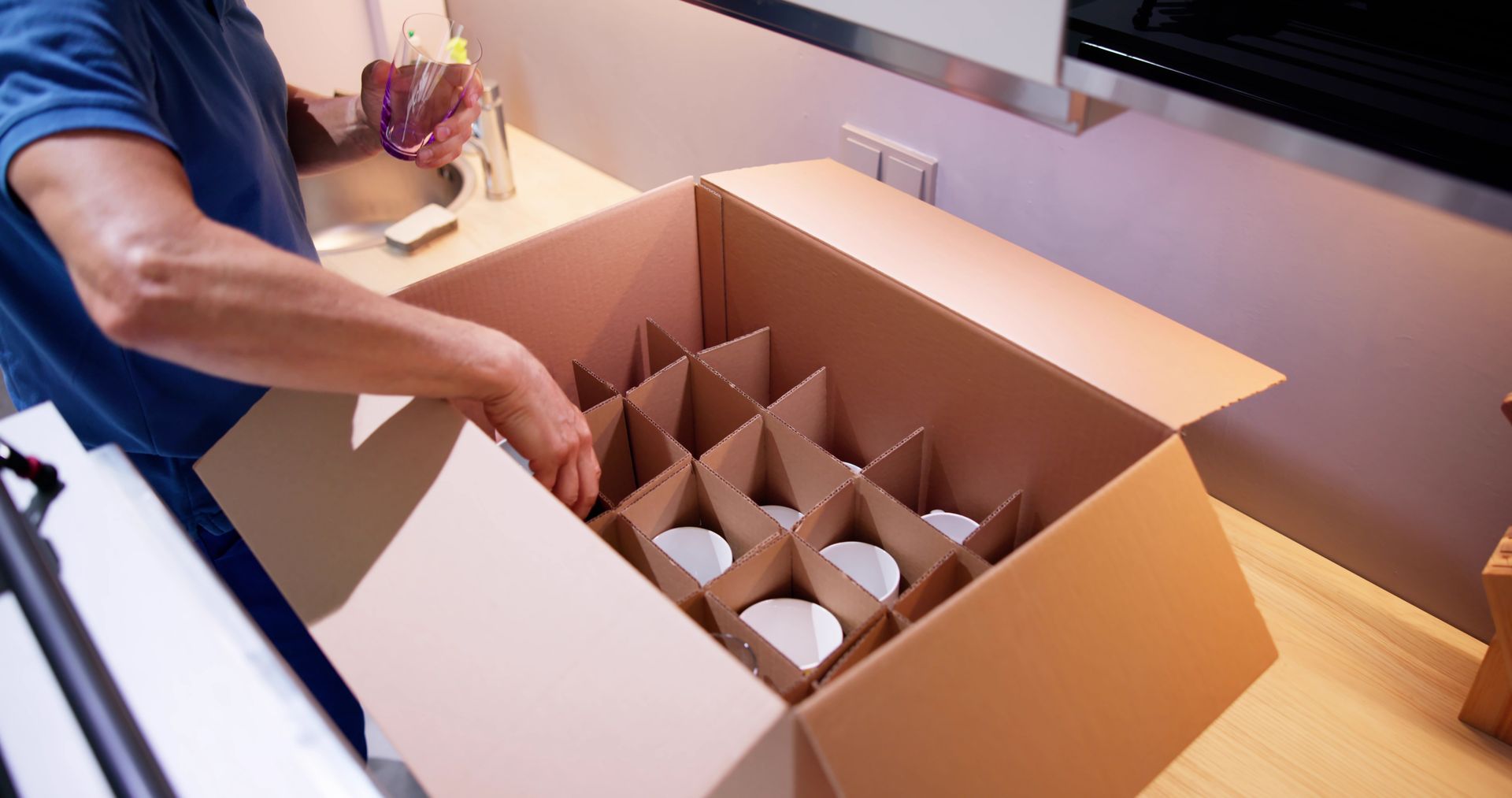 Person packing white cups into a cardboard box with dividers in a kitchen.