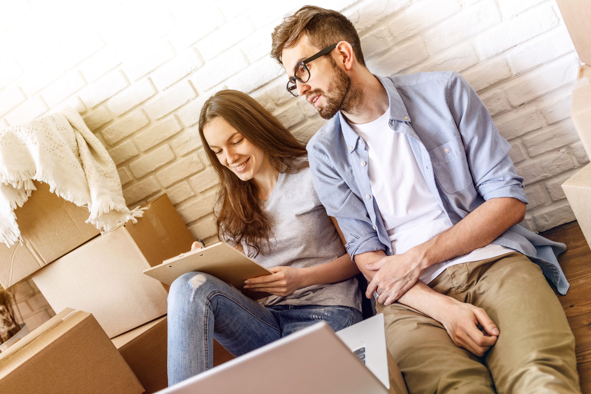 A couple sits surrounded by moving boxes, woman with clipboard, man looking on, smiling.