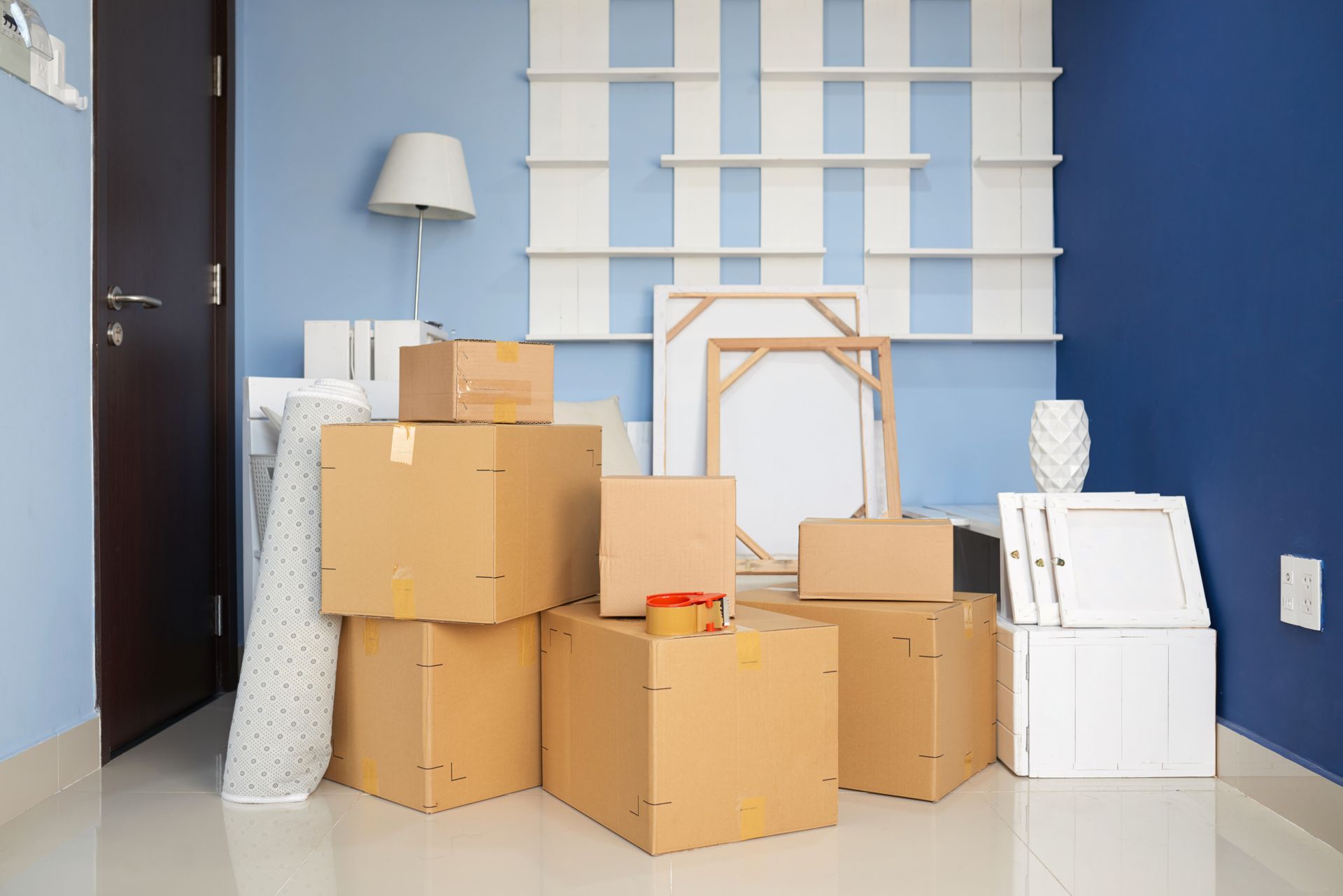 Boxes stacked in a room, ready for moving. Blue walls, door, white shelves in the background.