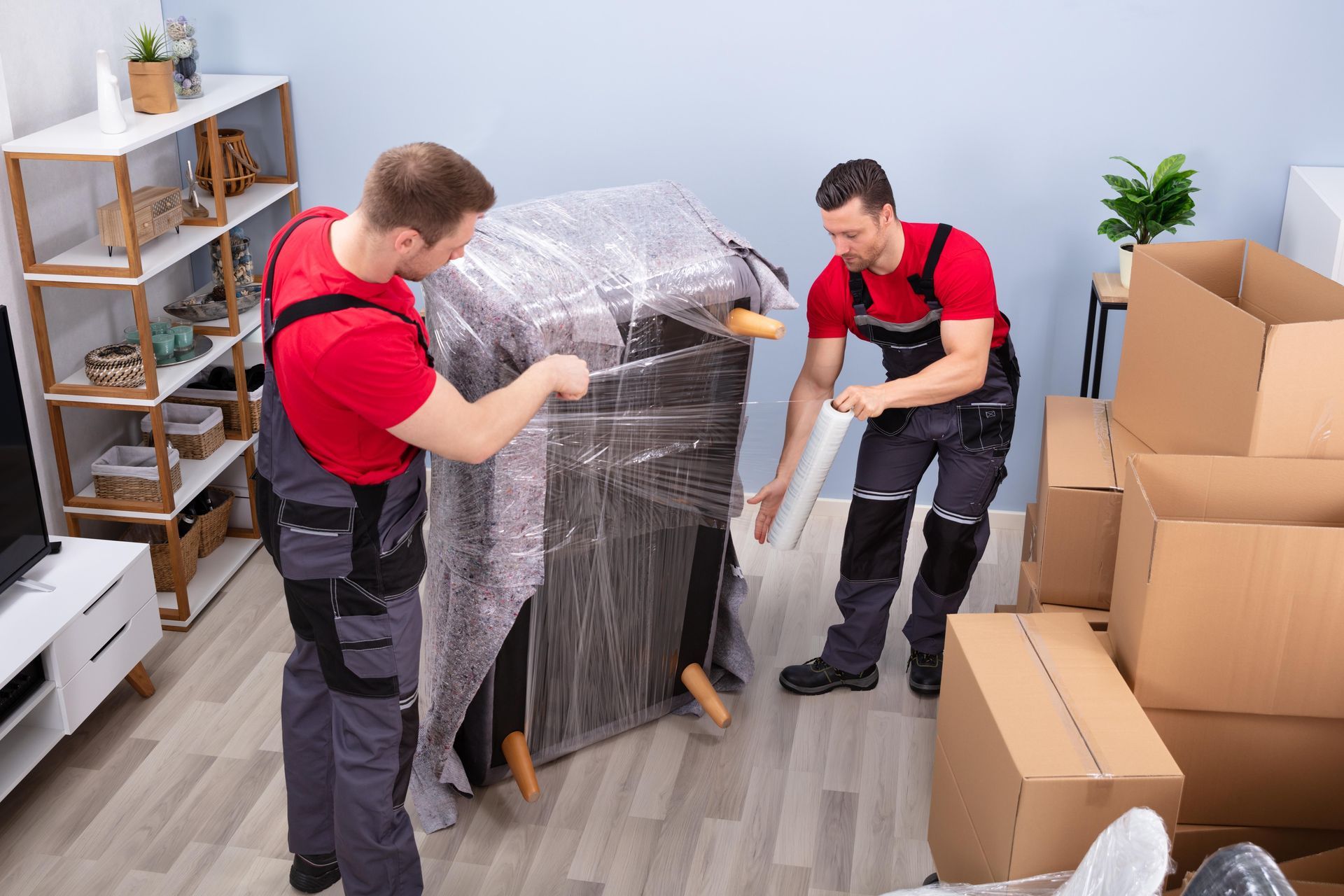 Two movers in red shirts wrapping furniture with plastic wrap near cardboard boxes.