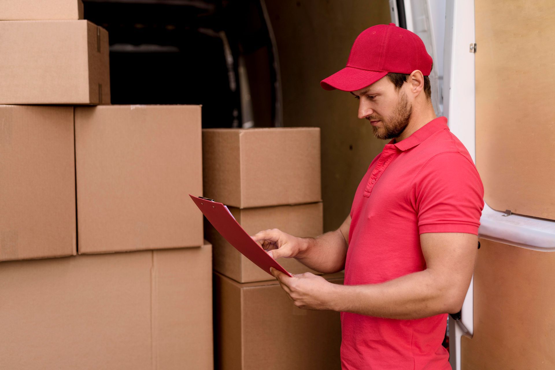 Delivery person in red uniform and cap, checks clipboard in front of packed van.