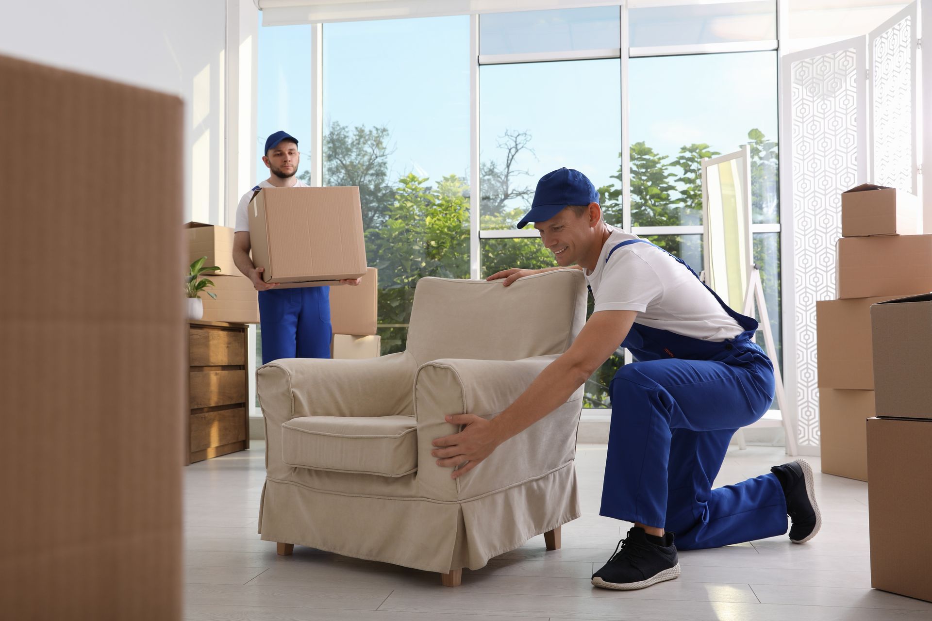 Movers packing furniture in a room with boxes. One kneels by a chair, while another holds a box.