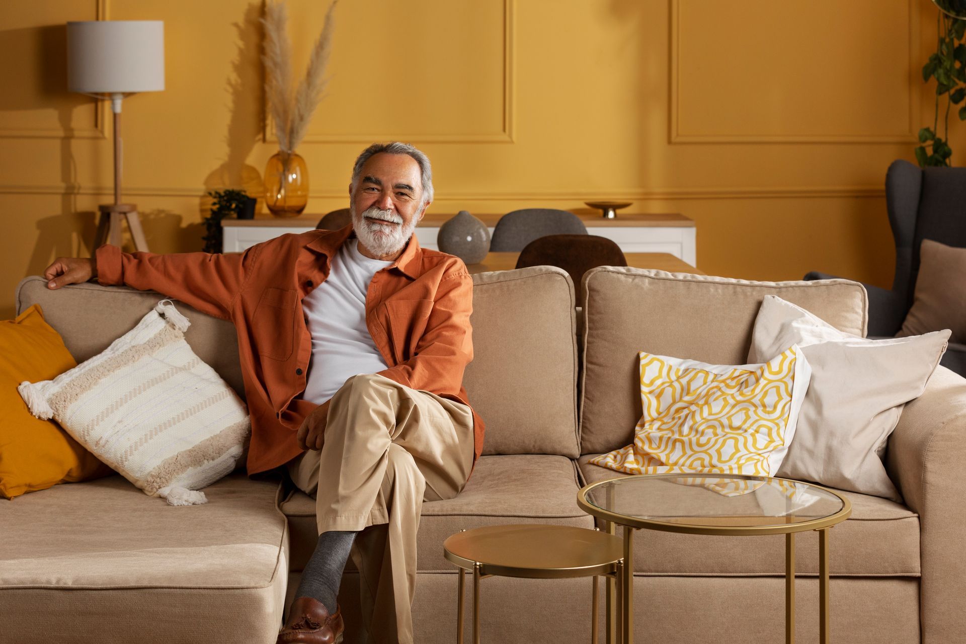 Man with gray beard sitting on a beige couch in a yellow-walled living room. He smiles, wearing an orange shirt and tan pants.