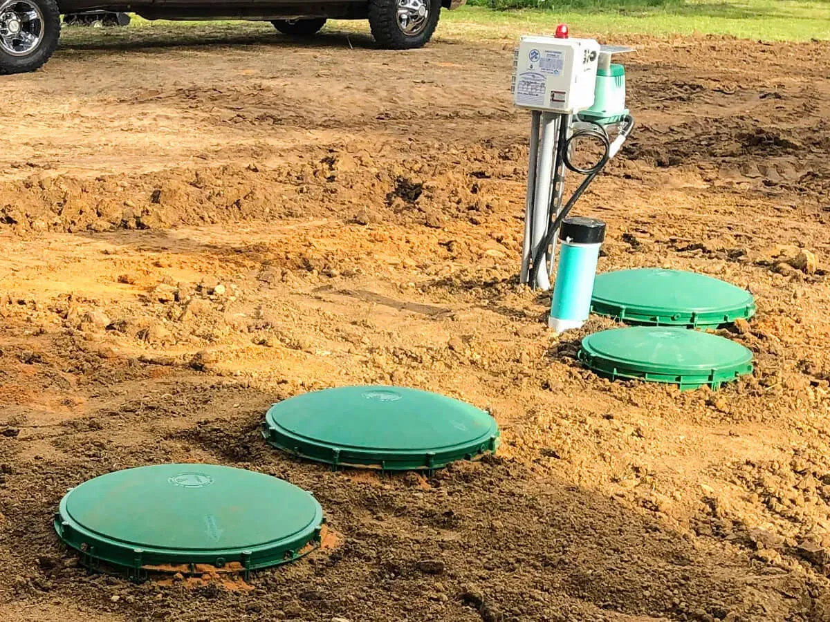 Green septic tank lids and control panel in a dirt yard. A truck is partially visible in the background.