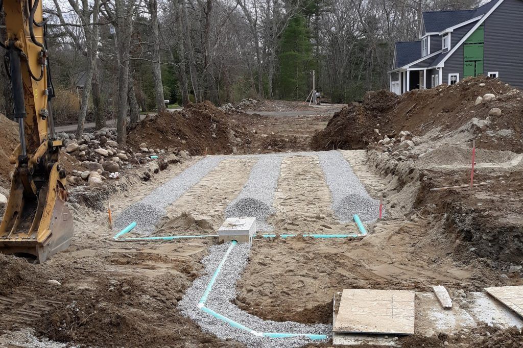 Construction site, septic system installation. Trenches with gravel, blue pipes, and heavy machinery in progress.