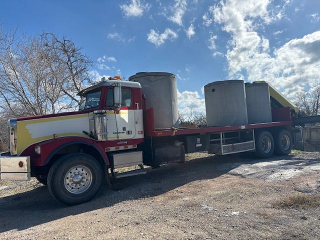 Red and white semi-truck carrying three large concrete cylinders on a flatbed trailer, under a blue sky.