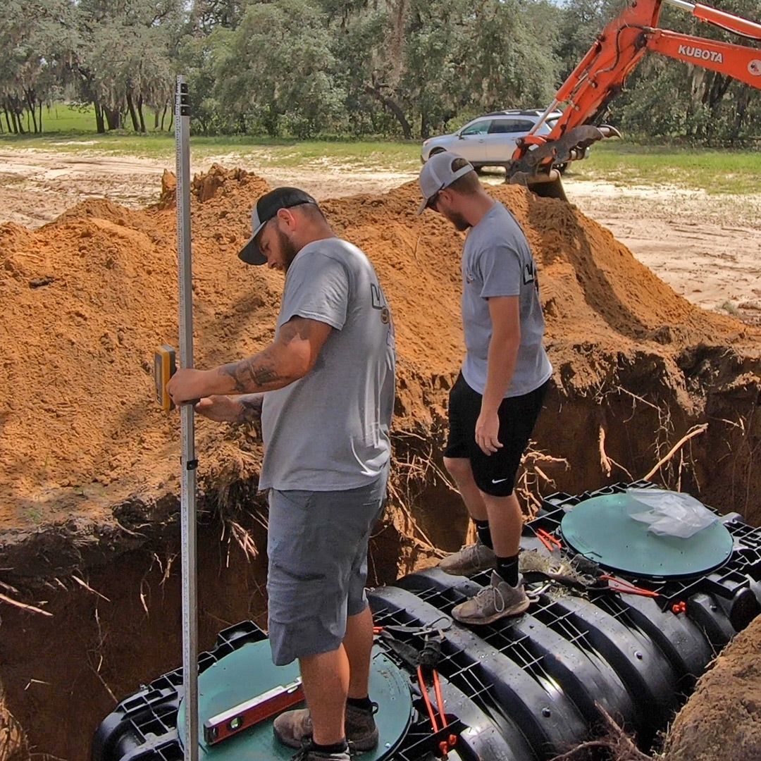 Two men installing septic tank in a construction site, one holding a level, the other checking the tank.