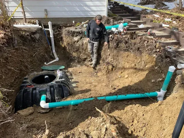 A person stands in an excavated area with septic tank and plumbing installation.