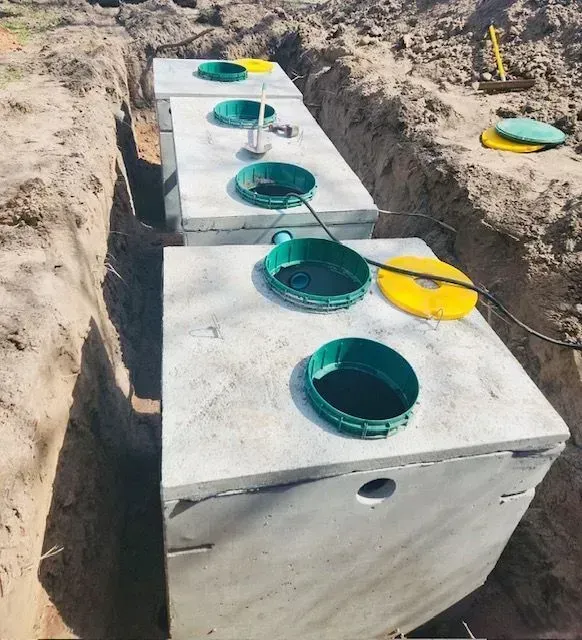Three concrete septic tanks in a trench; green and yellow access lids are open; dirt surrounds.
