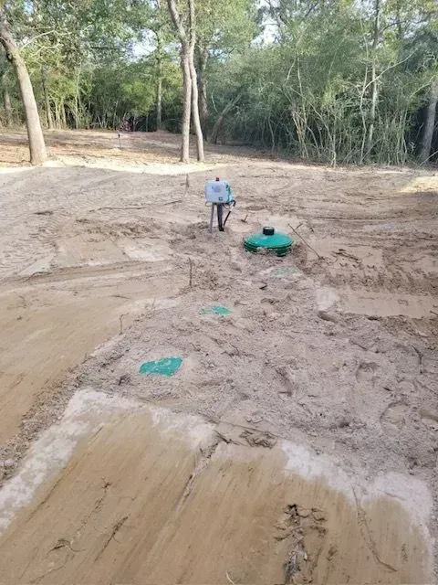 A person operating machinery on a dirt site with trees in the background, likely septic installation.