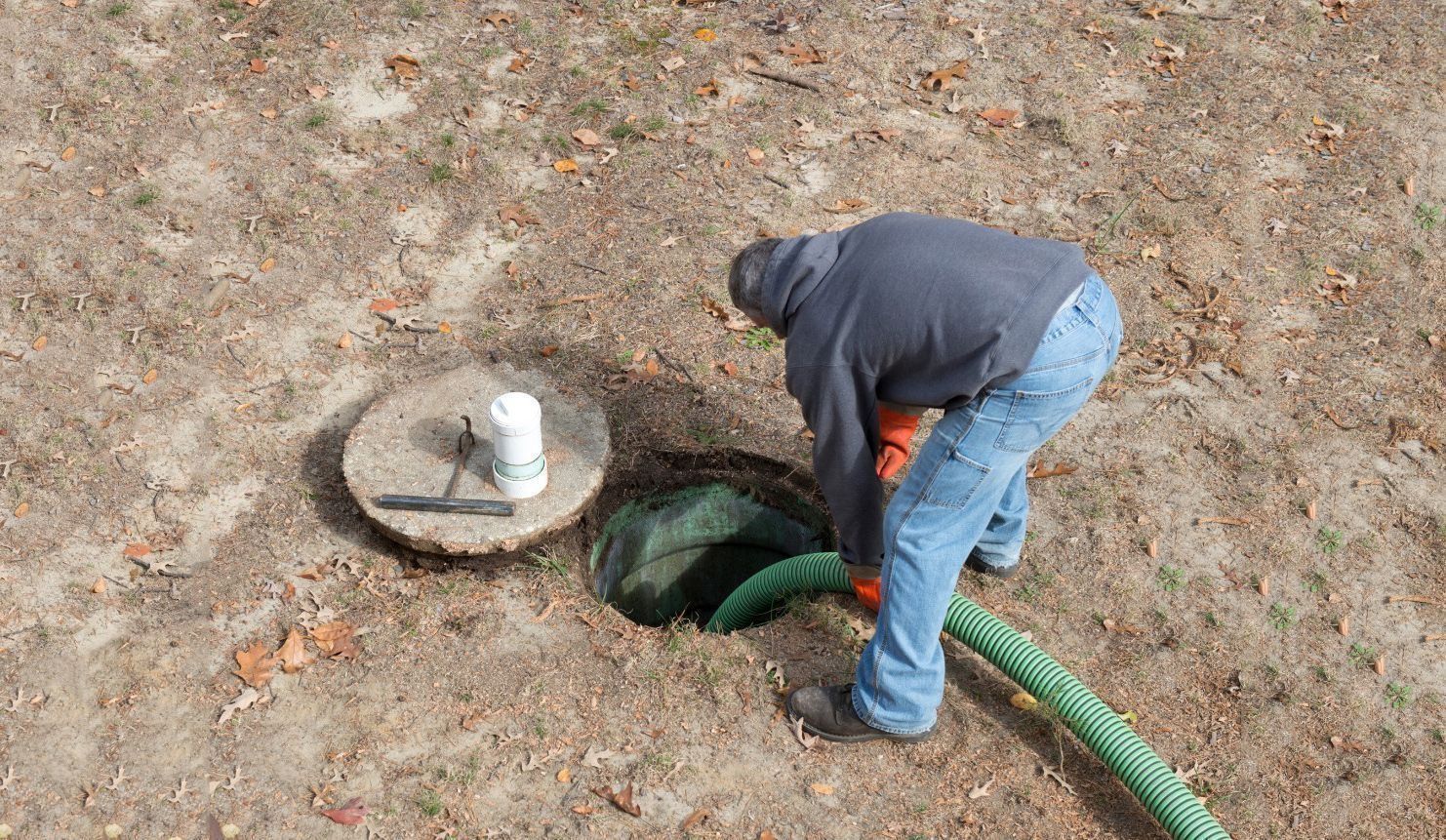 Man pumping a septic tank; outdoors. Person wears blue jeans and a gray shirt. Green hose and dirt.