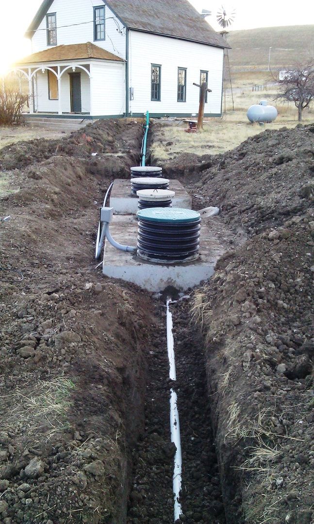 Construction of a septic system: trenches with pipes and tanks, house in background, outdoors.