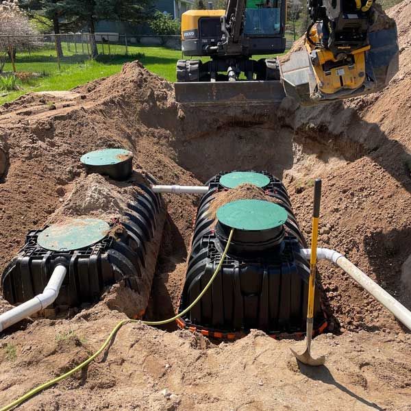 Two black septic tanks being installed in a dirt excavation, with machinery and a shovel.