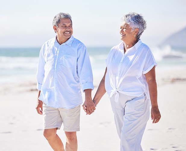 Couple holding hands, walking on a beach, smiling. White shirts, beige shorts, and a sunny day.
