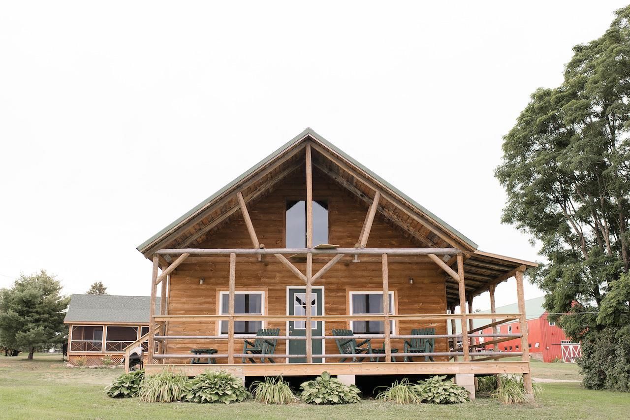 Rustic wooden cabin with a front porch and large windows, set in a grassy field with trees nearby