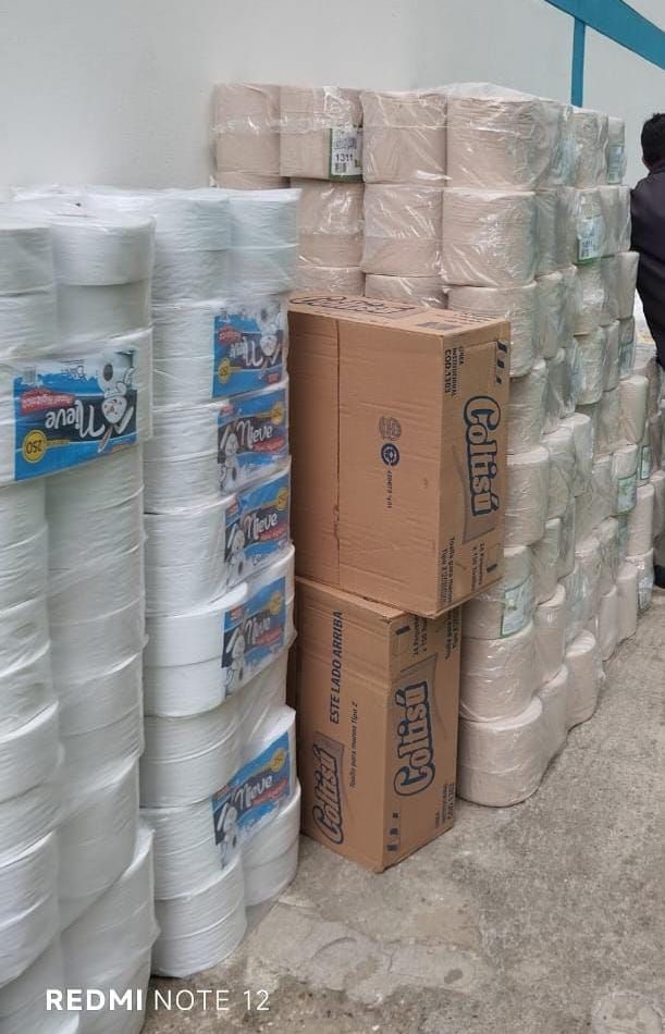 Pilas de cuerda blanca y rollos de papel envueltos cerca de cajas de cartón marrón. Persona al fondo.