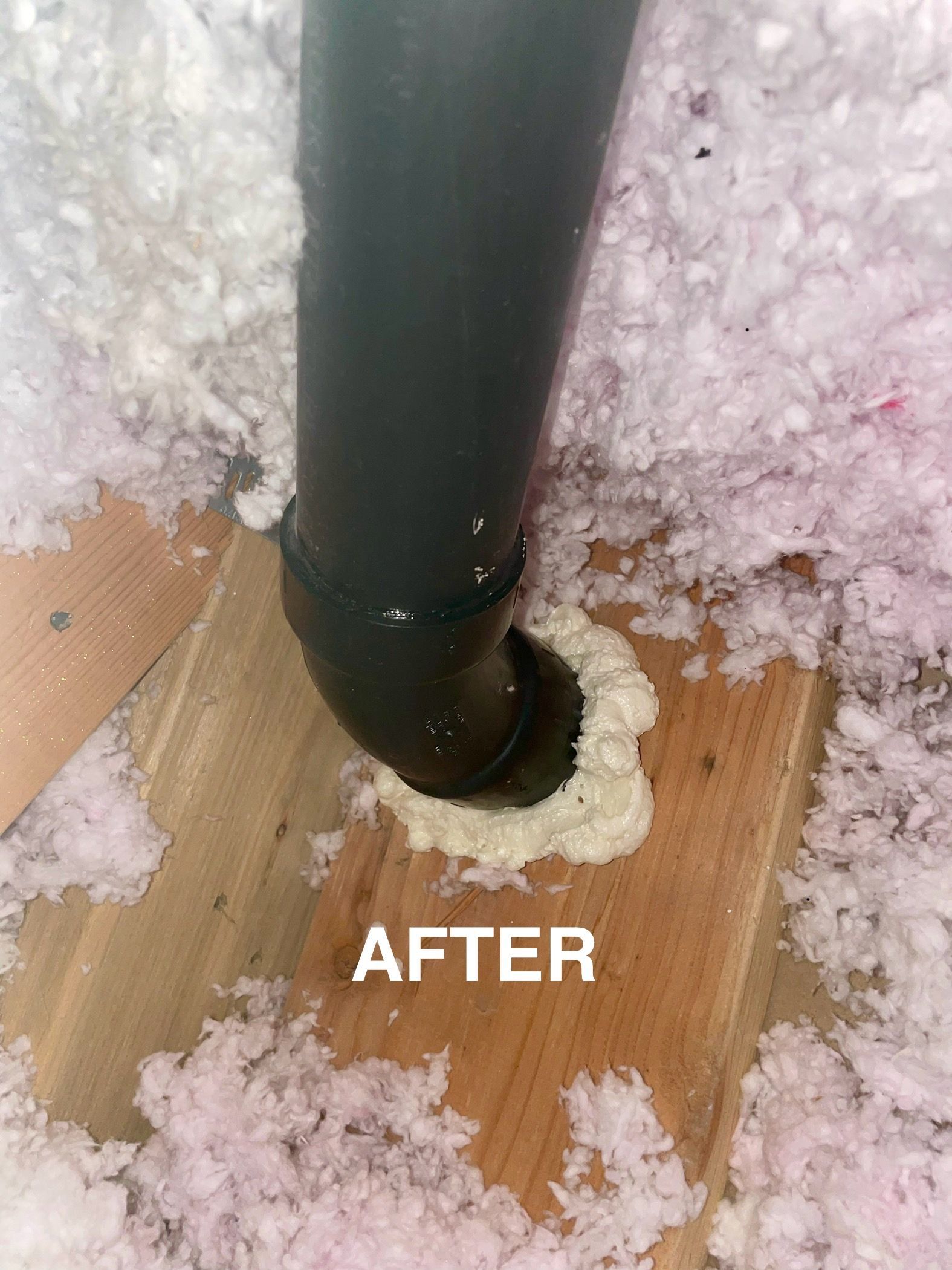 Black pipe sealed with expanding foam in an attic, surrounded by pink insulation and wood. 