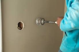 Person using a hole saw to cut a circular hole in a tan wall.