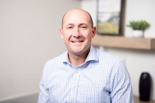 Joseph Hakes, MD in a light blue checkered shirt seated in a bright office setting