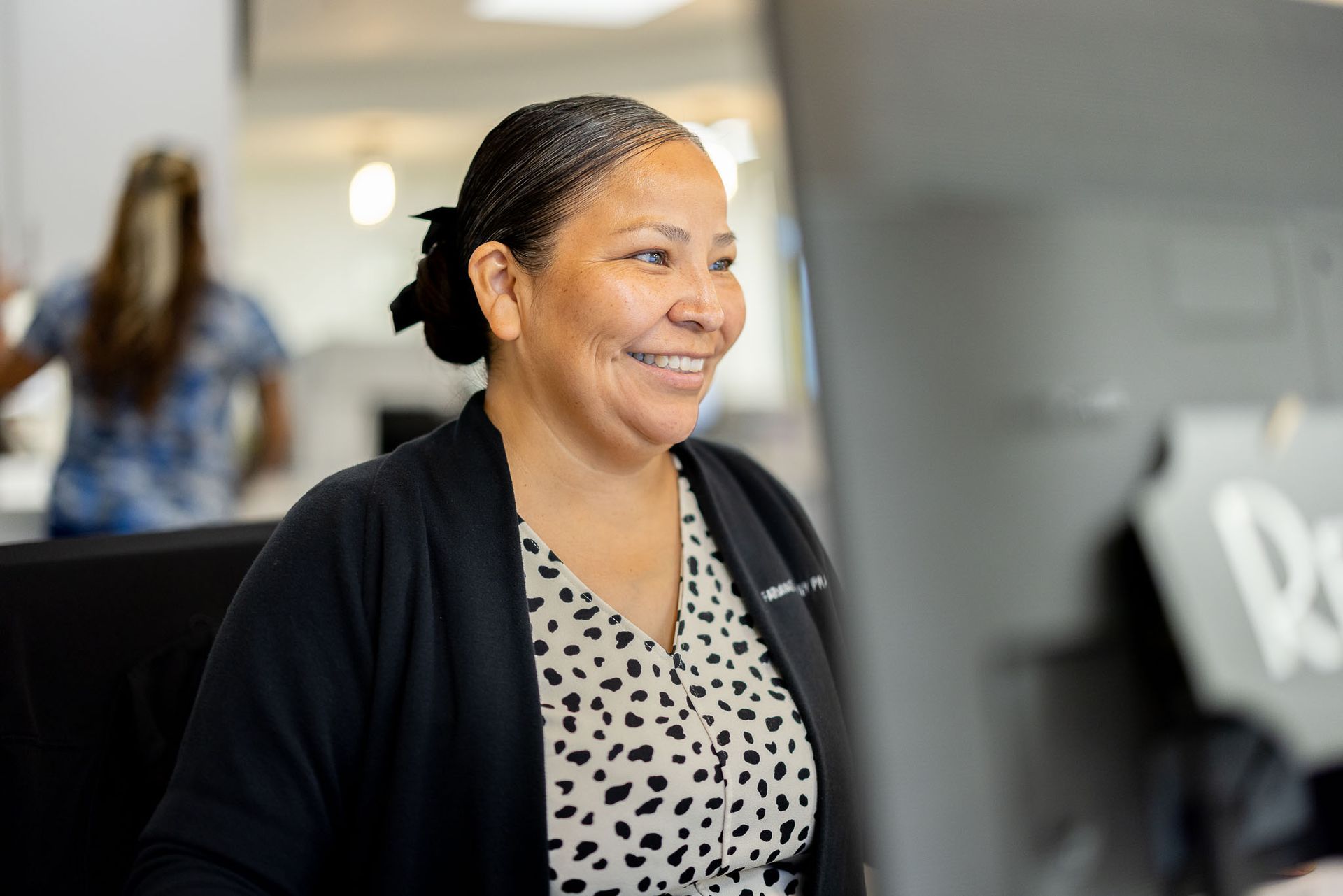 Smiling woman in a black cardigan stands in an office, looking at a computer screen.