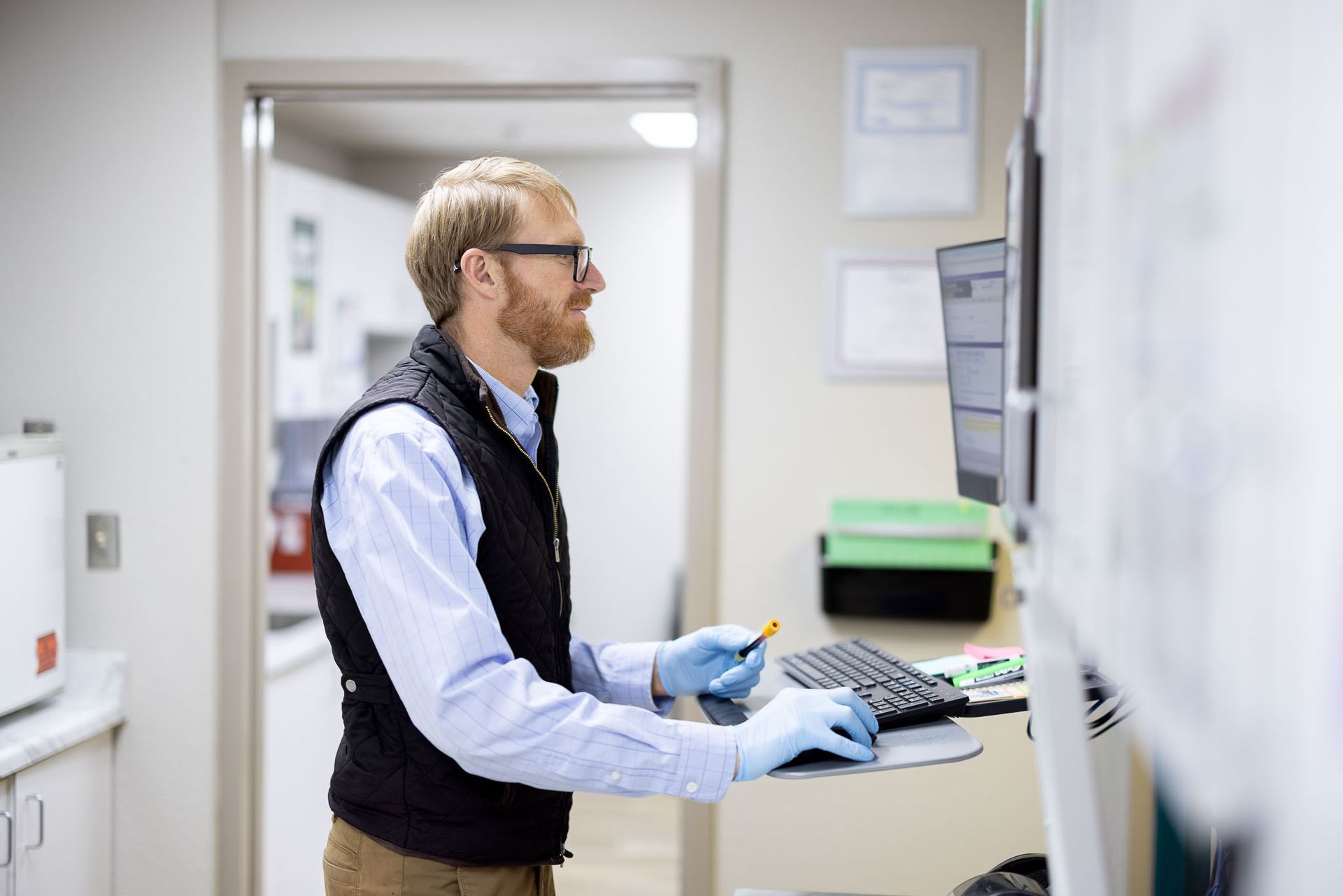 Person working at a computer station in an doctor's office, wearing blue gloves and a black vest.