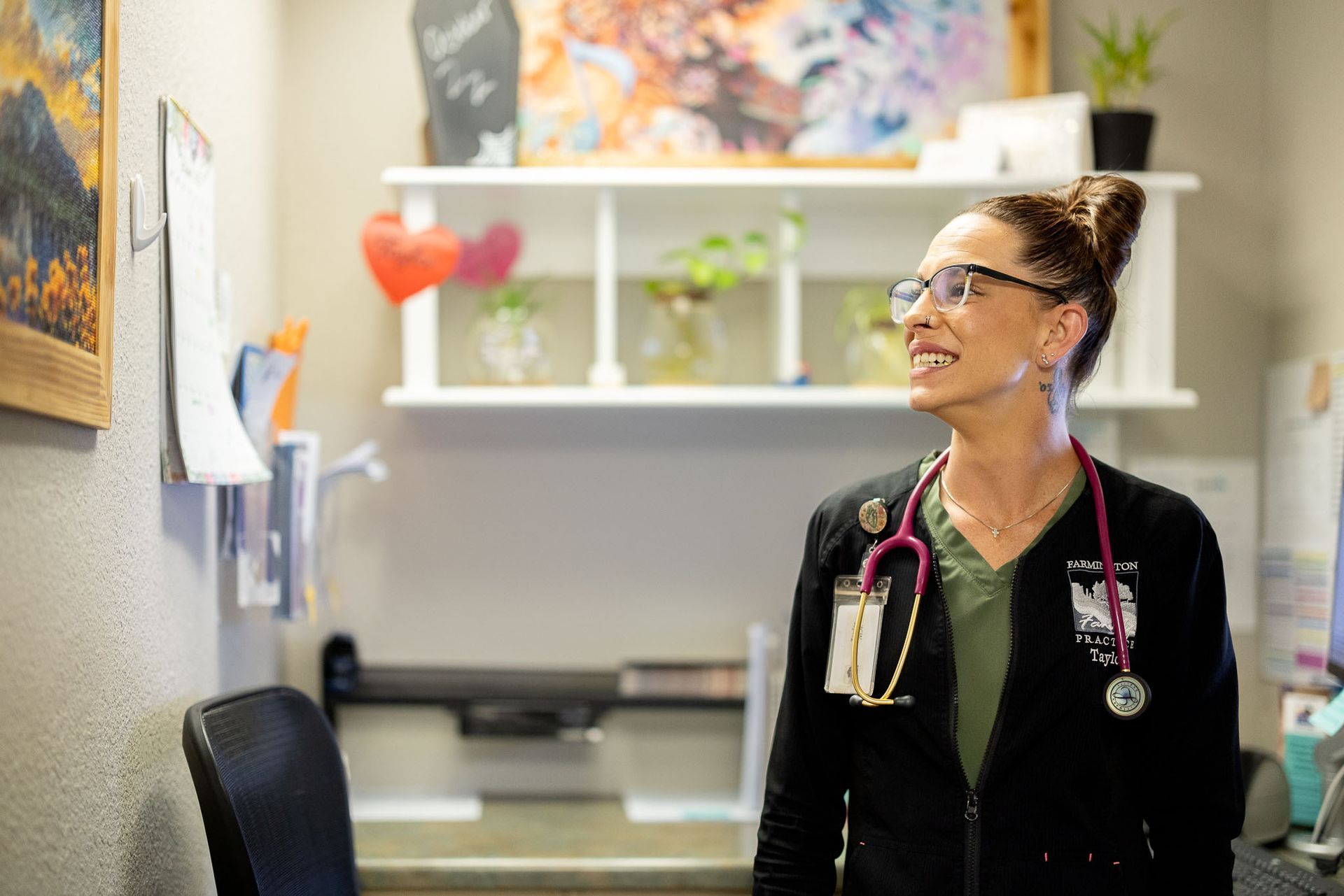 Nurse in black scrubs with stethoscope smiles in a clinic office, looking at a wall display.