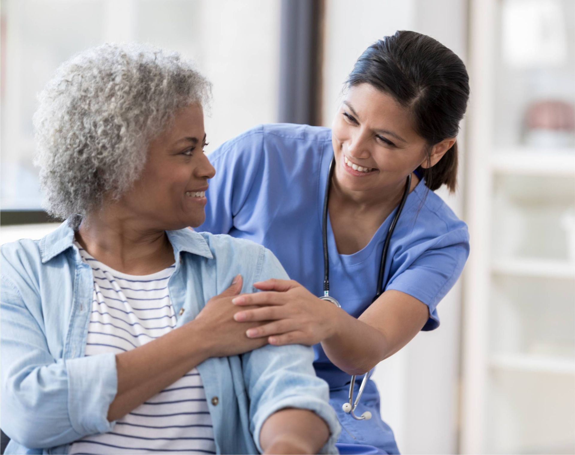 Nurse in blue scrubs gently comforts a smiling patient in a bright room