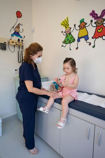 Nurse in blue scrubs helps a child seated on an exam table in a pediatric clinic with cartoon wall art.