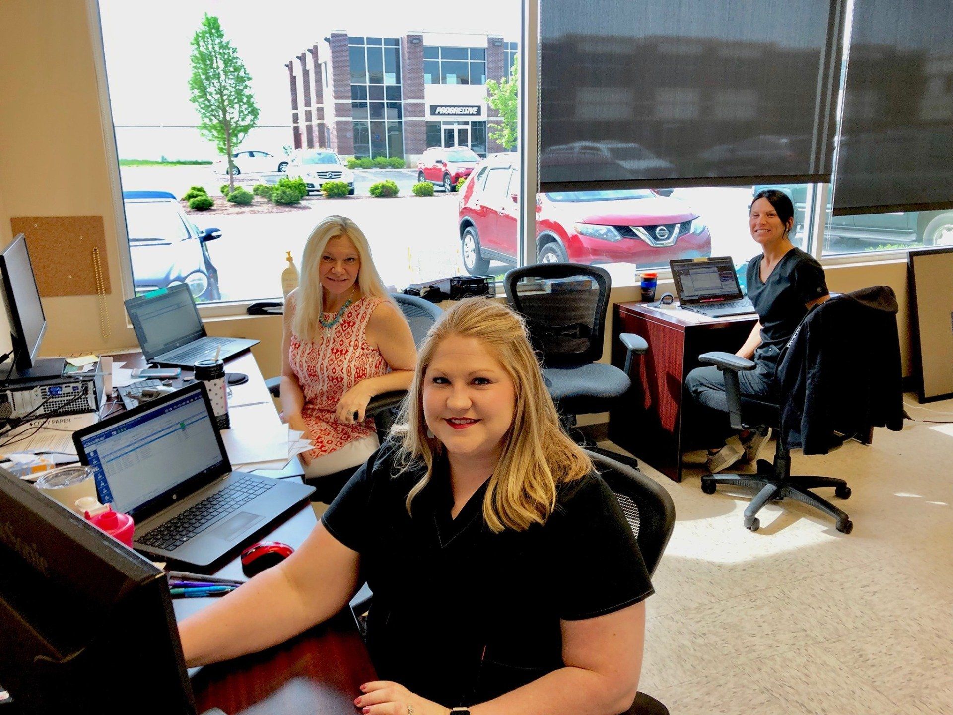 Three women are sitting at desks with laptops in an office.