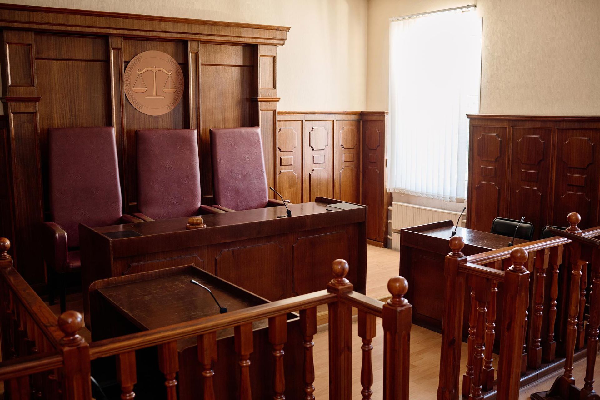 Courtroom with wooden judge's bench, chairs, and railing. Light from a window illuminates the room. 