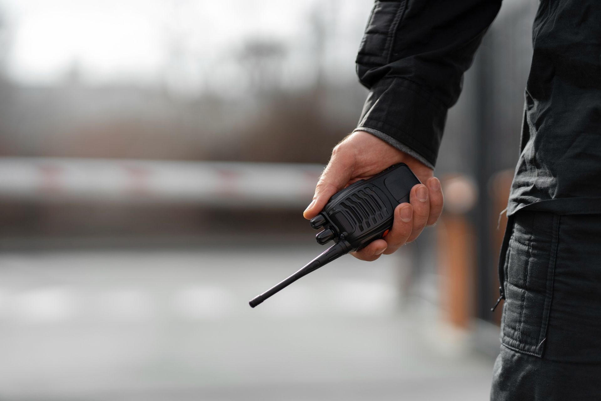 Man in black jacket holding a walkie-talkie in front of a gate. 