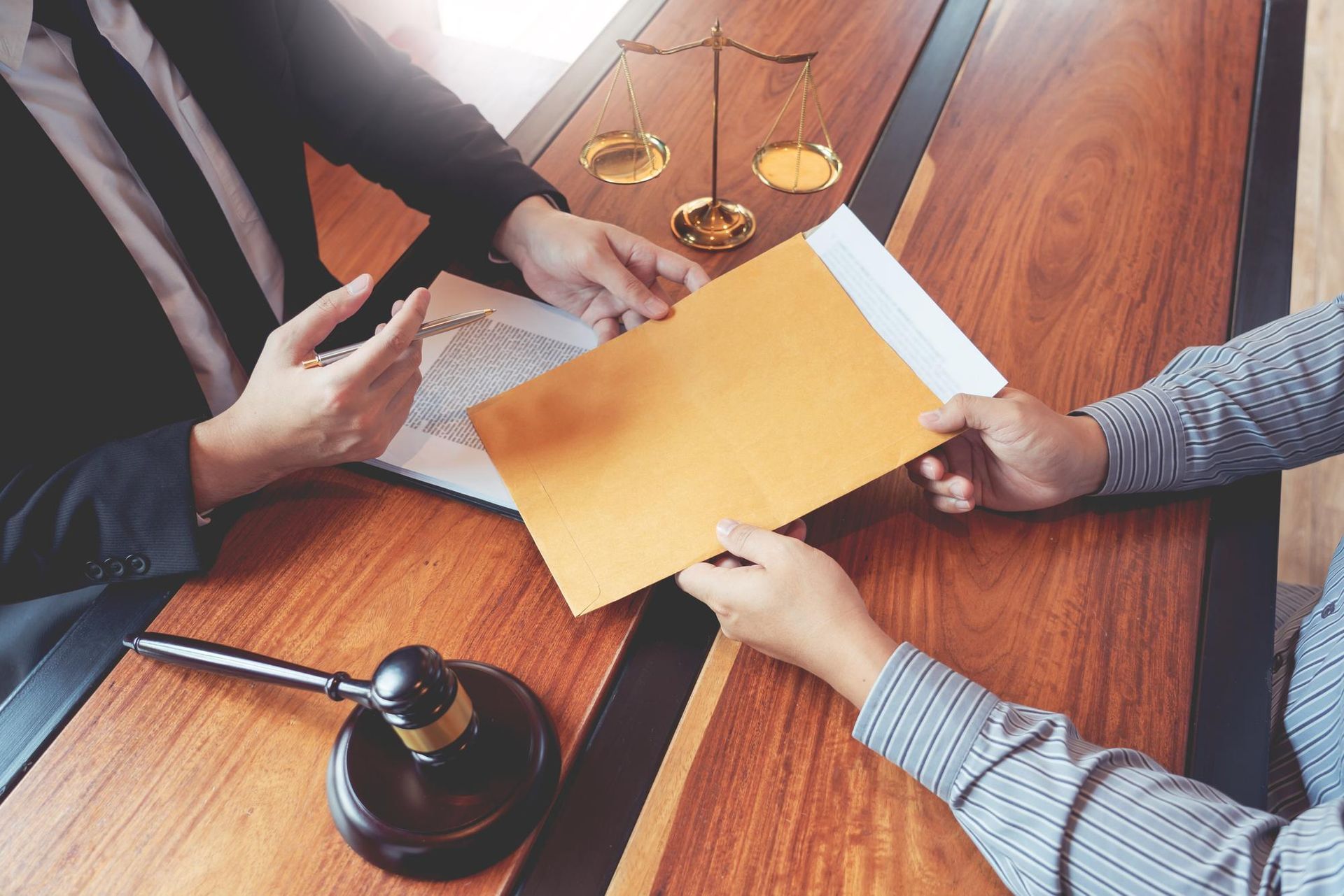Two people at a table; one hands an envelope. Scales of justice and gavel in view. 