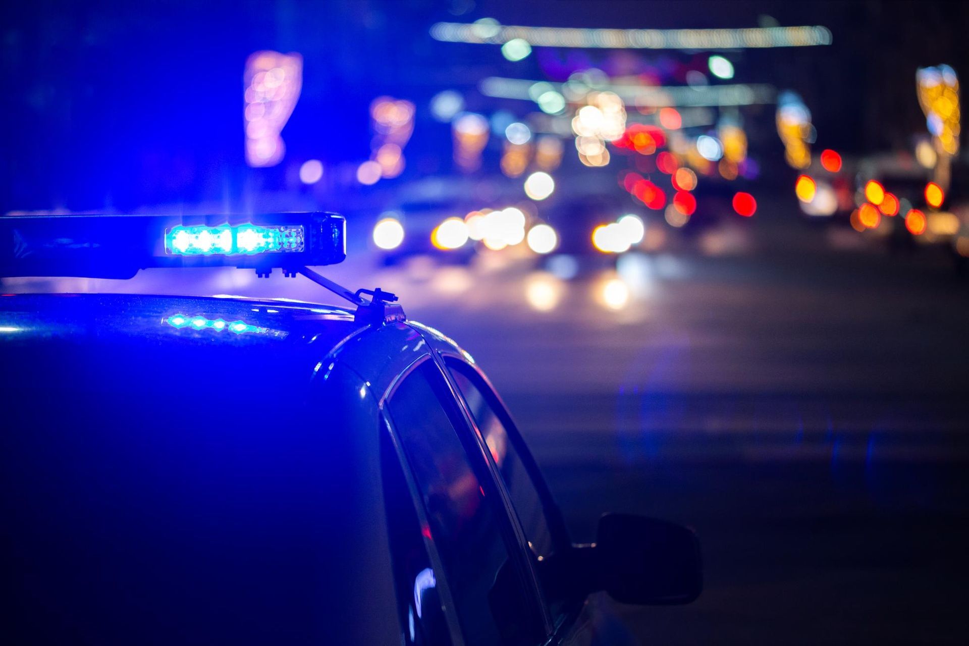 Police car with flashing blue lights on a city street at night, blurred traffic in background. 