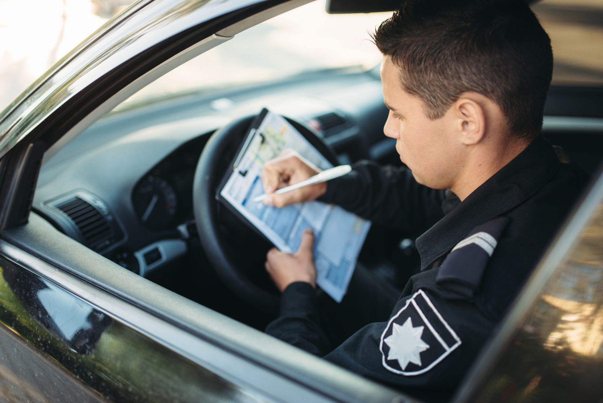 Police officer in uniform, writing on a notepad inside a black car. 