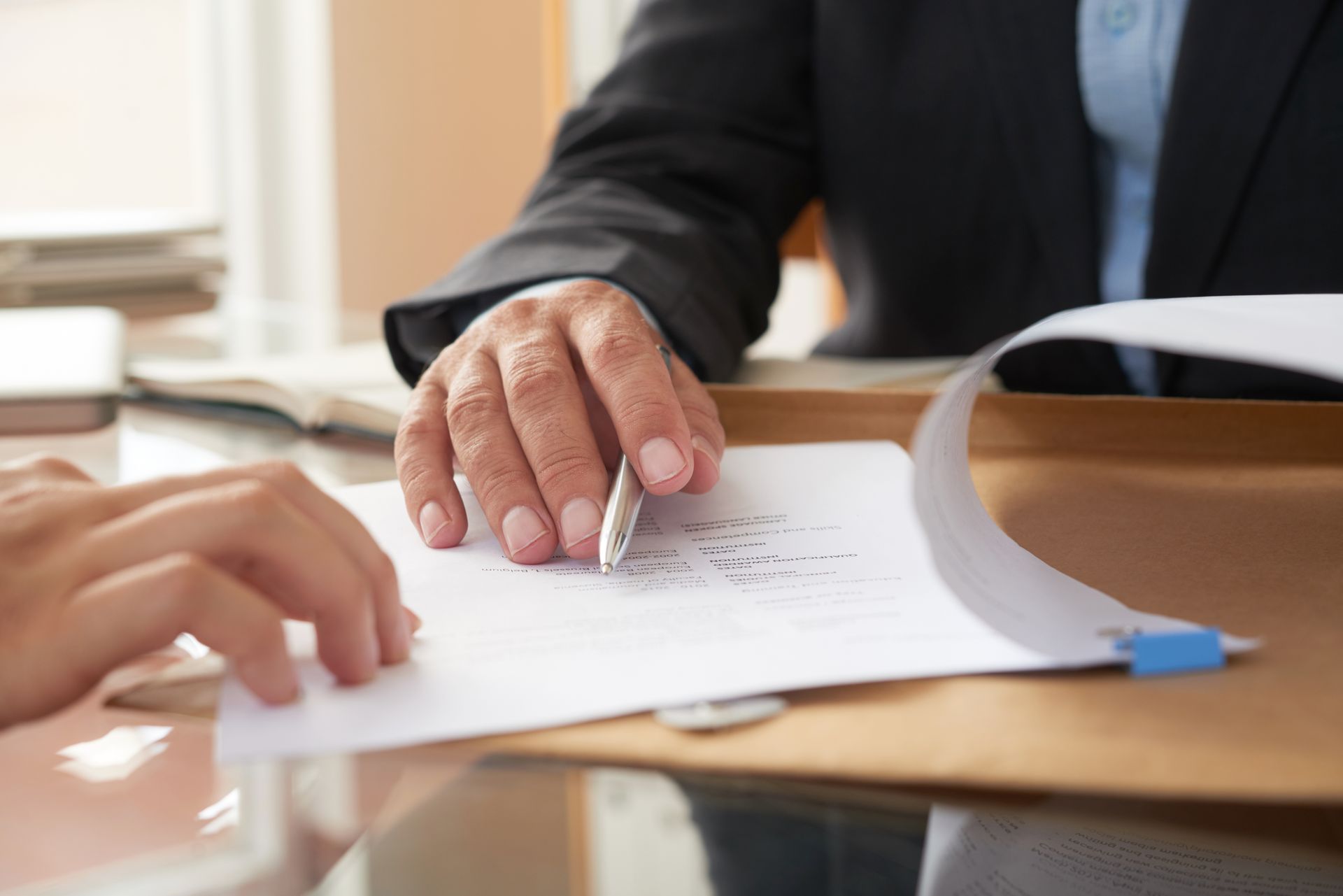 Hands over document; person in suit points pen at writing on paper. 