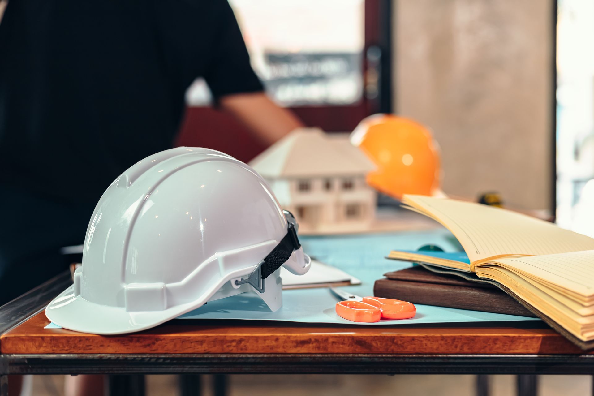 White hard hat on desk with blueprints, model house, and orange helmet; person in background.