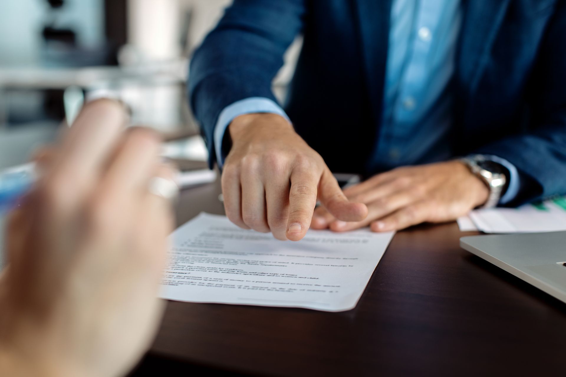 Person in blue suit pointing at document on a desk, another person's hand holding a pen.